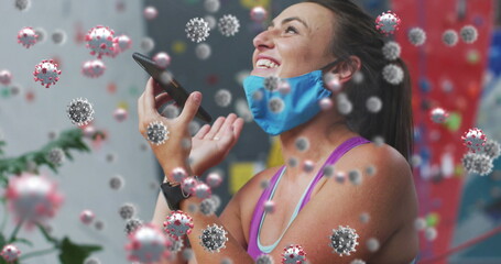 Smiling woman wearing blue cloth face mask and holding phone in rock-climbing gym, virus overlay