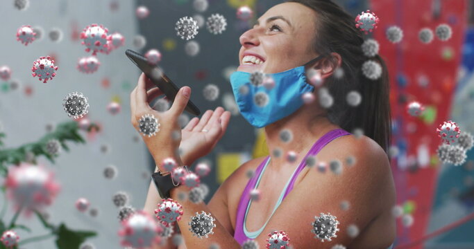 Smiling woman wearing blue cloth face mask and holding phone in rock-climbing gym, virus overlay - Powered by Adobe