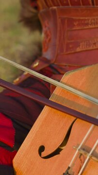 Musician in ethnic leather clothes plays igil fiddle-bow instrument sitting on green field closeup slow motion. Representation of old Yakut music