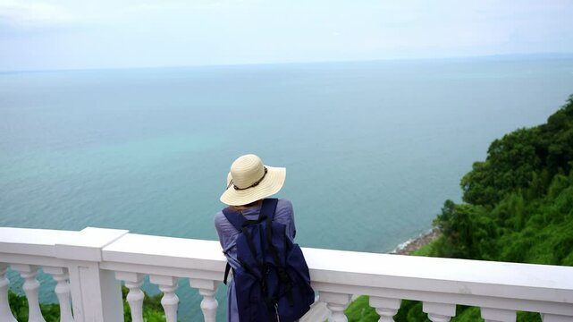 Female traveler with backpack and hat stopped at viewpoint looking at turquoise water, sandy coastal beach surrounded by trees, enjoying view. Concept of vacation, tourist destination, dreams, travel