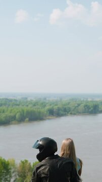 Biker in helmet hugs blonde girlfriend sitting together on motorcycle on green hill top near tranquil lake on spring day backside view slow motion