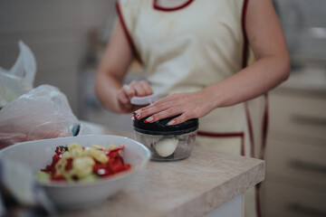 A woman uses a kitchen chopper to prepare a fresh salad with chopped vegetables at home, wearing an apron in a cozy kitchen setting. Focus is on her hands and the chopper.