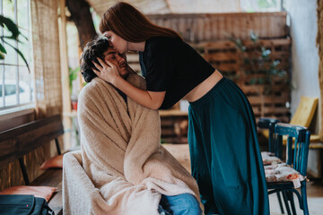 Fototapeta premium A caring woman gently kisses a man wrapped in a blanket, offering comfort in a warm setting. The scene exudes warmth, affection, and support, highlighting a tender moment of care.