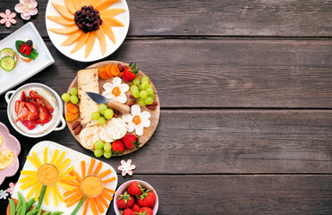Summer garden party food side border. Variety of fun flower theme charcuterie items, cheese, fruit, vegetables and finger snacks. Top view on a dark wood background.