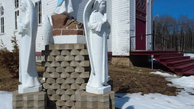 Monument composed of statues of the Virgin Mary with clasped hands and a trio of angels praying together near a church, rising to the top of the bell tower. Sainte-Florence, Quebec, Canada, 2025.