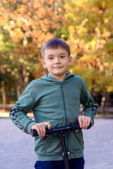 Happy boy in green hoodie riding scooter on sunny day in park. Green and yellow leaves in...