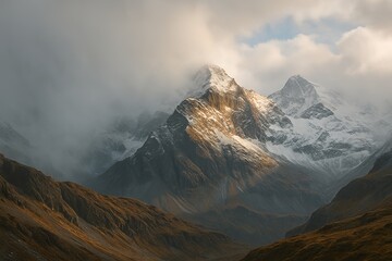 Fototapeta premium A breathtaking view of rugged mountain peaks dusted with snow, emerging from a valley of golden-brown terrain, partially shrouded in misty clouds as dramatic lighting breaks through
