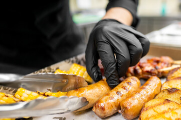 A chef with black gloves carefully arranges grilled sausages and corn on a serving tray, highlighting a delicious barbecue spread in a professional kitchen.
