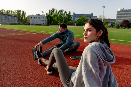Two athletes stretching their legs while seated on a running track in a stadium, focusing on flexibility and preparation for training