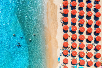 Aerial view of colorful red umbrellas on sandy beach, swimming people in blue sea on sunny summer...