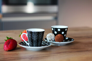 Two cups of coffee and saucers sit on a wooden table with strawberries and chocolate truffles. One cup is white with black floral designs, the other black with white polka dots.