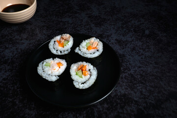 A plate of sushi on a black plate and a bowl of soy sauce. The sushi is rolled up and has a variety of ingredients including cucumber and carrots