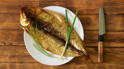 Top-view of smoked Baikal omul on a white plate with green onions and a wooden-handled knife, ideal...