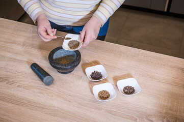 A woman's hand pours spices into a marble mortar. In the bowls are the spices: black pepper, zira and mustard seeds.