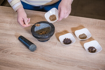 A woman's hand pours spices into a marble mortar. In the bowls are the spices: black pepper, zira and mustard seeds.