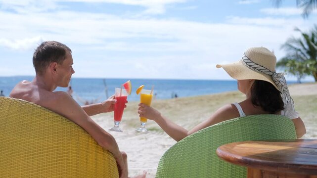 Sipping fresh fruit shakes, a young woman in a straw hat and a young man relaxing in beach chairs. They clink glasses, sharing a joyful moment on a scenic tropical shore, slow motion