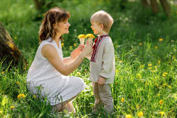 Young mother kneeling in the grass, smiling and playing with her little son who holds dandelion...