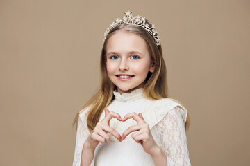 Portrait of a cheerful young girl in a white dress and tiara, making a heart shape with her hands...