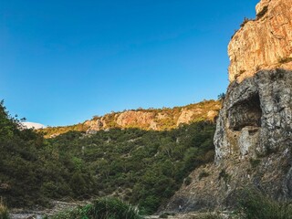 Sunlit rock cliff with a prominent cave opening, surrounded by greenery and contrasting against a vivid blue sky