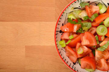 A partially visible colorful bowl of fresh tomato salad with green onions and basil sits on a wooden surface