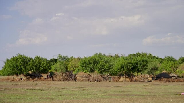 Safari-style village with thatched huts nestled in green bush. Grassy plains stretch under soft blue sky with scattered clouds. Dense green trees line the horizon, offering natural shelter