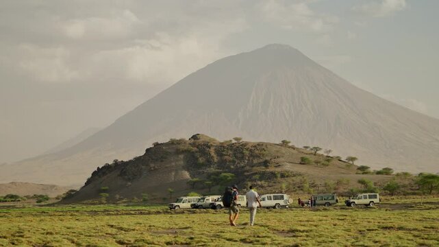 Tourists and guides with 4x4 vehicles by Ol Doinyo Lengai volcano in Tanzania. People visiting the famous natural park in Africa with raw of safari cars and trucks by hill with mountain in background