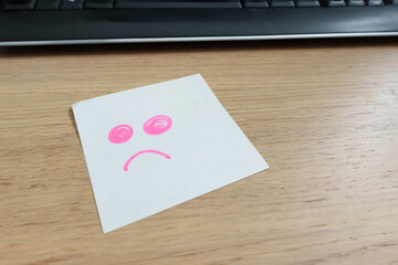 Sad pink face drawn in a white note by placing on a wooden desktop with keyboard. Sticky notes