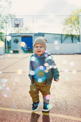 Toddler boy running outside with soap bubbles. Child on a warm spring day for a walk in a jacket and hat.