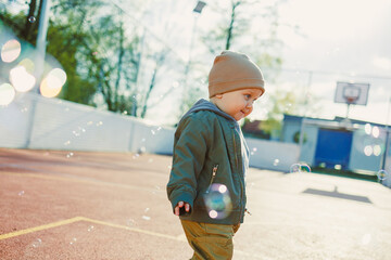 Cheerful 2 year old boy playing outside in summer in a green jacket with soap bubbles. Toddler boy running outside and playing with bubbles.