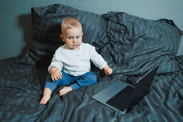 A 2-year-old boy sits in bed and presses buttons on a laptop. The child plays with a laptop.