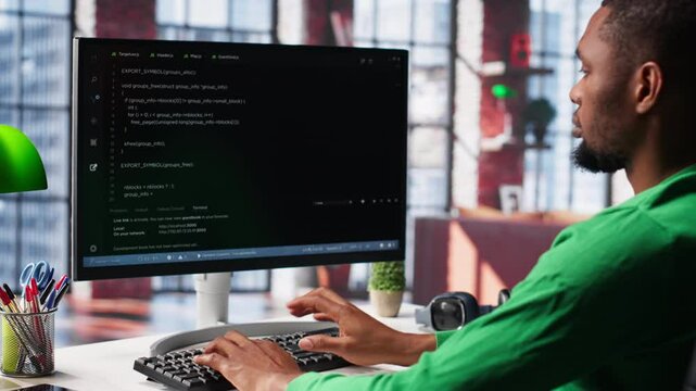Male programmer types code on computer and running script, programming language. African american IT expert in modern loft fixing errors within the software, implementing new features. Camera A.