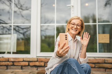 Modern mature woman sit at garden use mobile phone talk on video call