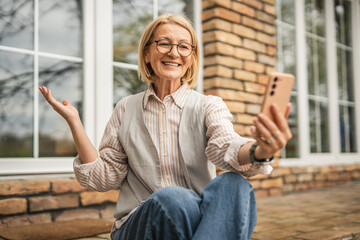 Modern mature woman sit at garden use mobile phone talk on video call