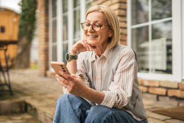 Modern casual mature woman sit at garden use mobile phone and enjoy