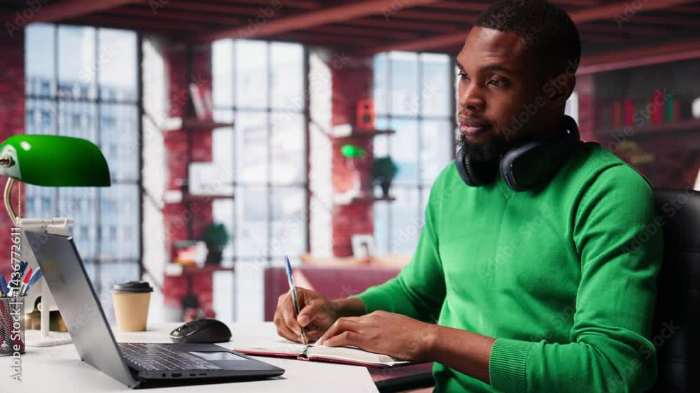 Poster smiling black man taking notes while watching a tutorial presentation, doing online self learning an