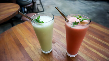 Two refreshing fruit smoothies in tall glasses with metal straws on a wooden table, served in a cozy outdoor café with a green natural background