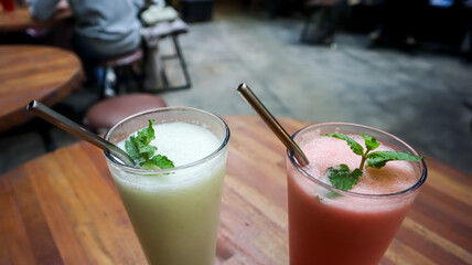 Two refreshing fruit smoothies in tall glasses with metal straws on a wooden table, served in a cozy outdoor café with a green natural background