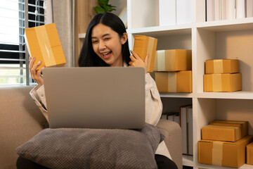 A woman is holding two boxes and smiling. She is sitting in front of a laptop. The boxes are brown and appear to be gifts. The woman seems happy and excited about the boxes
