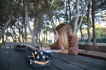 Girl Writes Music Notes On Sheet Music Sitting At The Park Table In The Nature