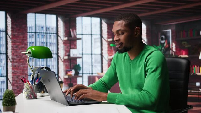 African american remote worker solving his daily freelancing tasks on laptop, working in relaxation from the comfort of his modern loft. Tech savvy black man internet user, multitasking. Camera B.