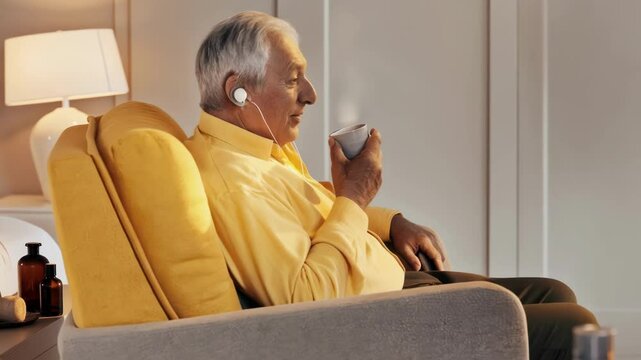 An older gentleman in a cozy armchair surrounded by wellness paraphernalia holds a cup of herbal tea listening attentively through his wireless earbuds to advice shared by fellow