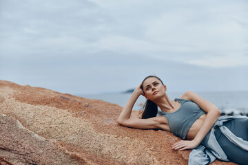 Fototapeta premium Young woman in activewear lounging on a rocky beach, showcasing a serene expression and relaxed posture, perfect for wellness and fitness themes