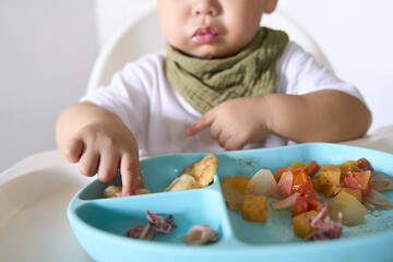 15 month old baby is sitting on high chair eating with fingers