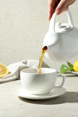 Woman pouring aromatic tea from teapot into cup at light textured table, closeup