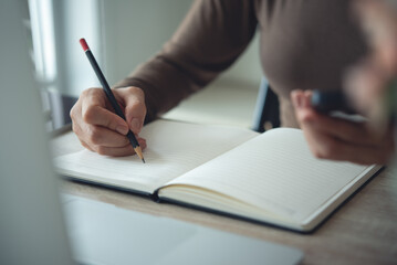 Close up, Woman hand with a pencil writing on paper notebook with laptop computer on office table. Business woman taking note on notebook, planning work schedule. Student study online, e-learning