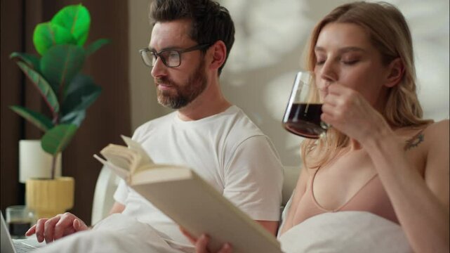 Busy Caucasian pair woman reading book in bed female drink coffee morning routine in bedroom man working boyfriend browsing laptop online web male typing gadget couple girlfriend relax speaking family