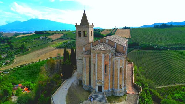 OFFIDA, ITALY - AUGUST 4, 2024: Aerial fly-over push-in shot of Chiesa di Santa Maria della Rocca. Group poses for photos in front of the church while stunning blue mountains rise in the distance