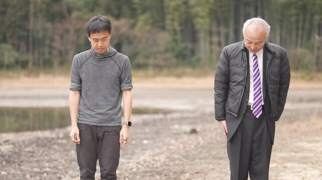 In a bamboo grove and pond park, a man in his 70s in a suit and a man in his 30s practice tai chi. Their slow movements are captured in a slow-motion video against the peaceful natural background.