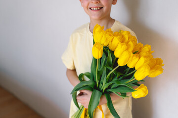 A boy stands indoors holding a large bouquet of yellow tulips, radiating happiness with a big smile on his face. The bright atmosphere reflects cheerfulness and warmth. Mother's Day and spring concept