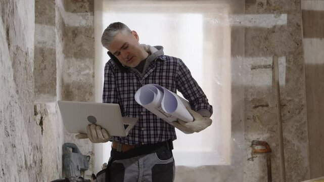 Confused puzzled construction builder worker on a renovation building site using laptop and talking on phone reading house blueprints and trying to understand the project layout, tasks and work to do